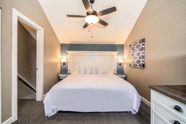 A bedroom with a large bed, white bedding, two nightstands, wall lights, a ceiling fan, and geometric artwork on the wall. A doorway is on the left.