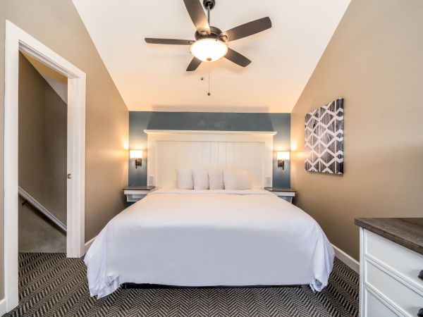 A bedroom with a large bed, white bedding, two nightstands, wall lights, a ceiling fan, and geometric artwork on the wall. A doorway is on the left.