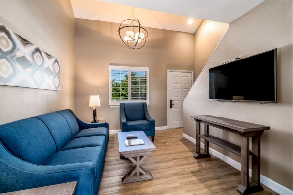 A cozy living room with blue furniture, a TV, a lamp, and a modern chandelier. Wooden flooring and wall art complete the look.