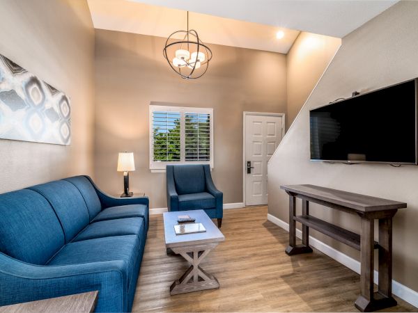 A cozy living room with blue furniture, a TV, a lamp, and a modern chandelier. Wooden flooring and wall art complete the look.