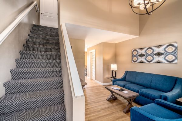 The image shows a cozy living room with a staircase, blue couch, wooden floor, and a patterned rug, featuring modern artwork on the wall.