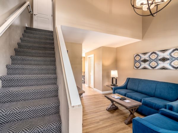 The image shows a cozy living room with a staircase, blue couch, wooden floor, and a patterned rug, featuring modern artwork on the wall.