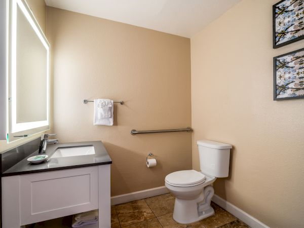 A bathroom with a sink, mirror, toilet, and towel racks, featuring two framed pictures on beige walls and tiled flooring.