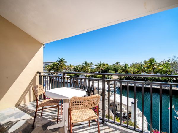 A balcony with a table and two chairs overlooks a waterway and lush palm trees under a clear blue sky.