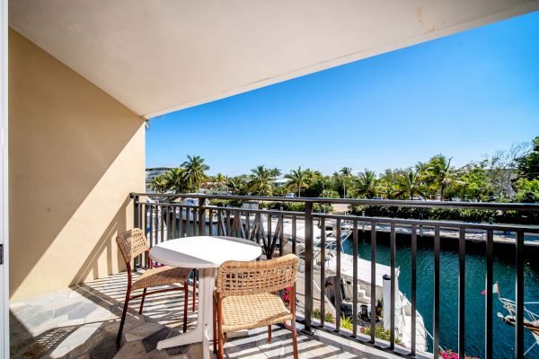 A balcony with two wicker chairs and a table overlooks a marina with boats, surrounded by palm trees under a clear blue sky.