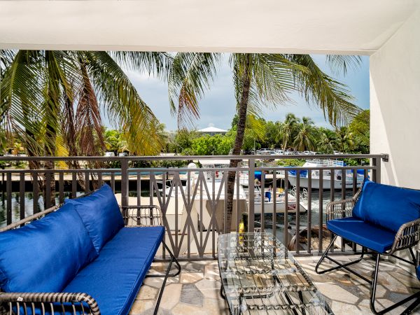 A cozy balcony with blue cushioned seating, glass table, palm trees, and sunshine in a tropical setting.