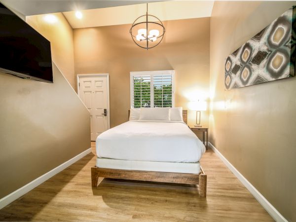A cozy modern bedroom with a wooden platform bed, beige walls, a wall-mounted TV, framed art, a ceiling light, a door and window at the far end, and hardwood floors.