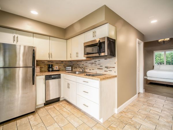 A modern L-shaped kitchen with stainless steel fridge, microwave, dishwasher, and light wood cabinets; tiled floor, beige walls, and a glimpse into a bedroom.