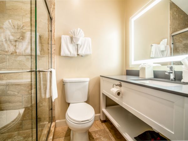 A modern bathroom with a glass-enclosed shower, a white toilet, a long vanity, and a backlit mirror, all in neutral tones.