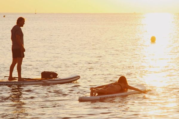 Two people on paddleboards enjoy a calm sunset on the water, one standing and one lying down, with gentle waves around them.