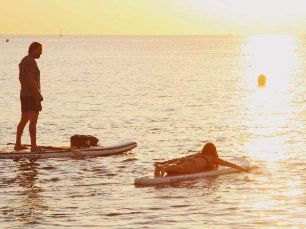 Two people on paddleboards enjoy a calm sunset on the water, one standing and one lying down, with gentle waves around them.