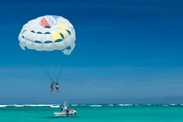 Two people parasailing over clear turquoise water, tethered to a boat under a bright blue sky.