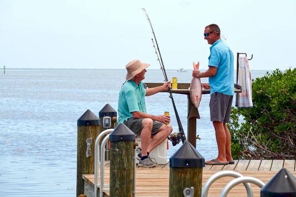 Two men on a dock, one holding a fish and a fishing rod, the other sitting with a drink. They're by the water on a clear day.