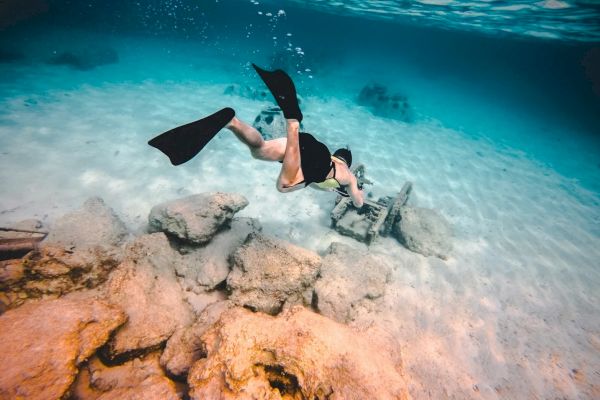 A snorkeler swims underwater over rocks and a submerged chair in clear blue water.