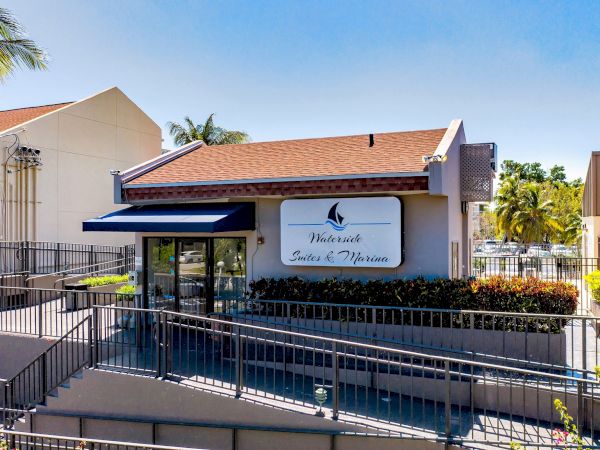 Exterior of Waterside Suites & Marina hotel with entrance sign, blue awning, railings, and palm trees under a clear sky.