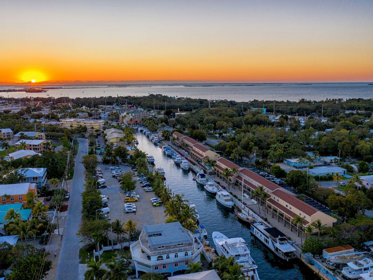 A marina at sunset with a calm canal, yachts docked along both sides, palm trees, and colorful buildings.