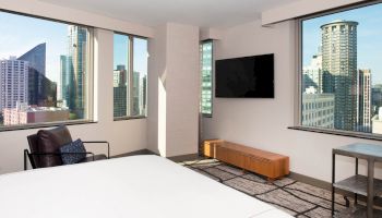 The image shows a modern hotel room with large windows offering city views, a mounted TV, a lounge chair, and simple contemporary furnishings.
