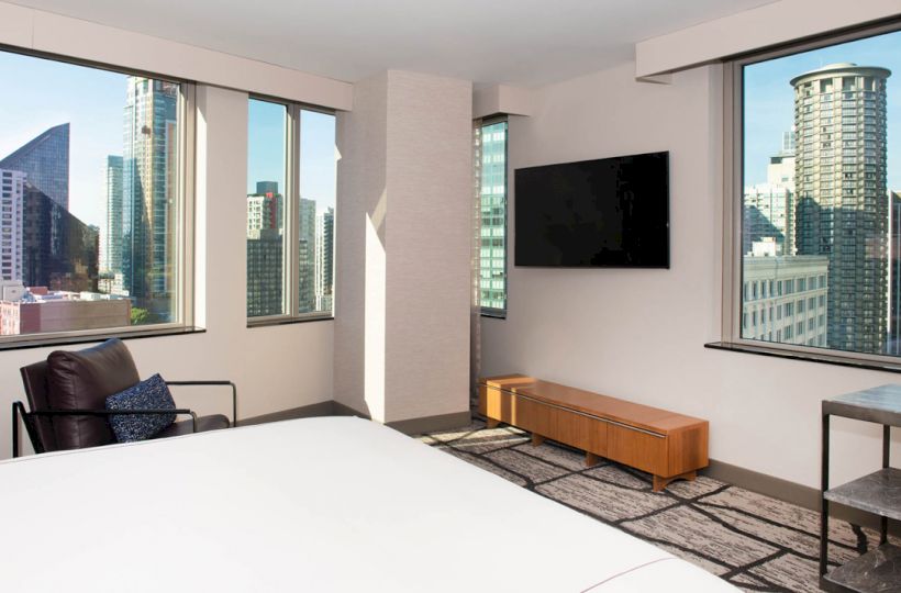 The image shows a modern hotel room with large windows offering city views, a mounted TV, a lounge chair, and simple contemporary furnishings.