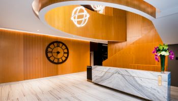 The image shows a modern reception area with a marble desk, wood-paneled walls, a large wall clock, and a decorative spiral staircase.