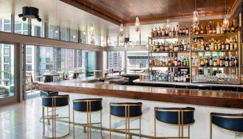 A modern bar with high shelves full of liquor bottles, stylish pendant lights, and blue swivel chairs is framed by large windows and city views.
