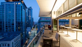 The image shows a modern cityscape at dusk from a high balcony that has tables and chairs, overlooking buildings and an illuminated street.