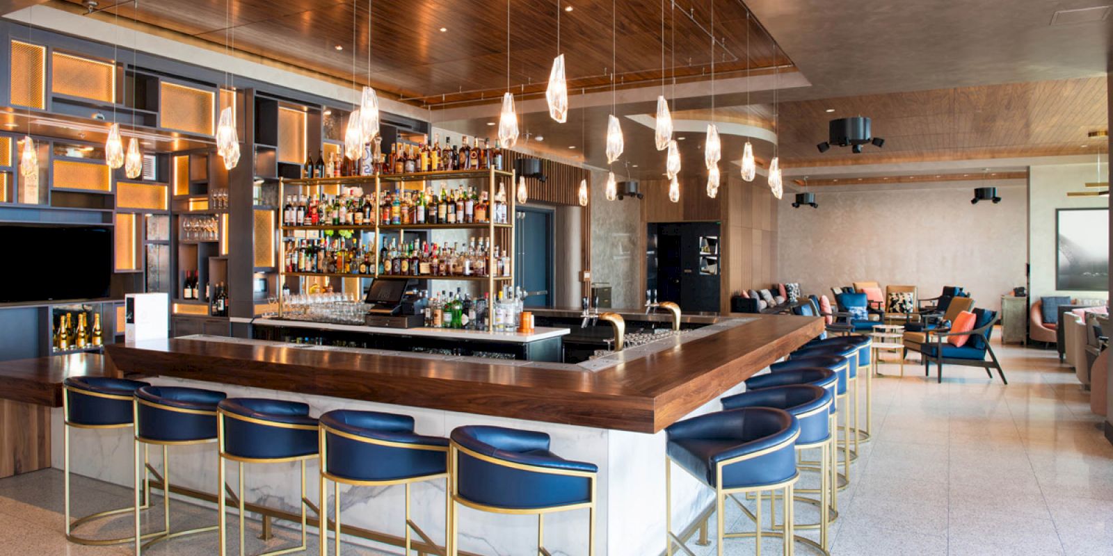 The image shows a modern bar with a wooden counter, high-end bar stools, various bottles behind the bar, and stylish hanging lights.