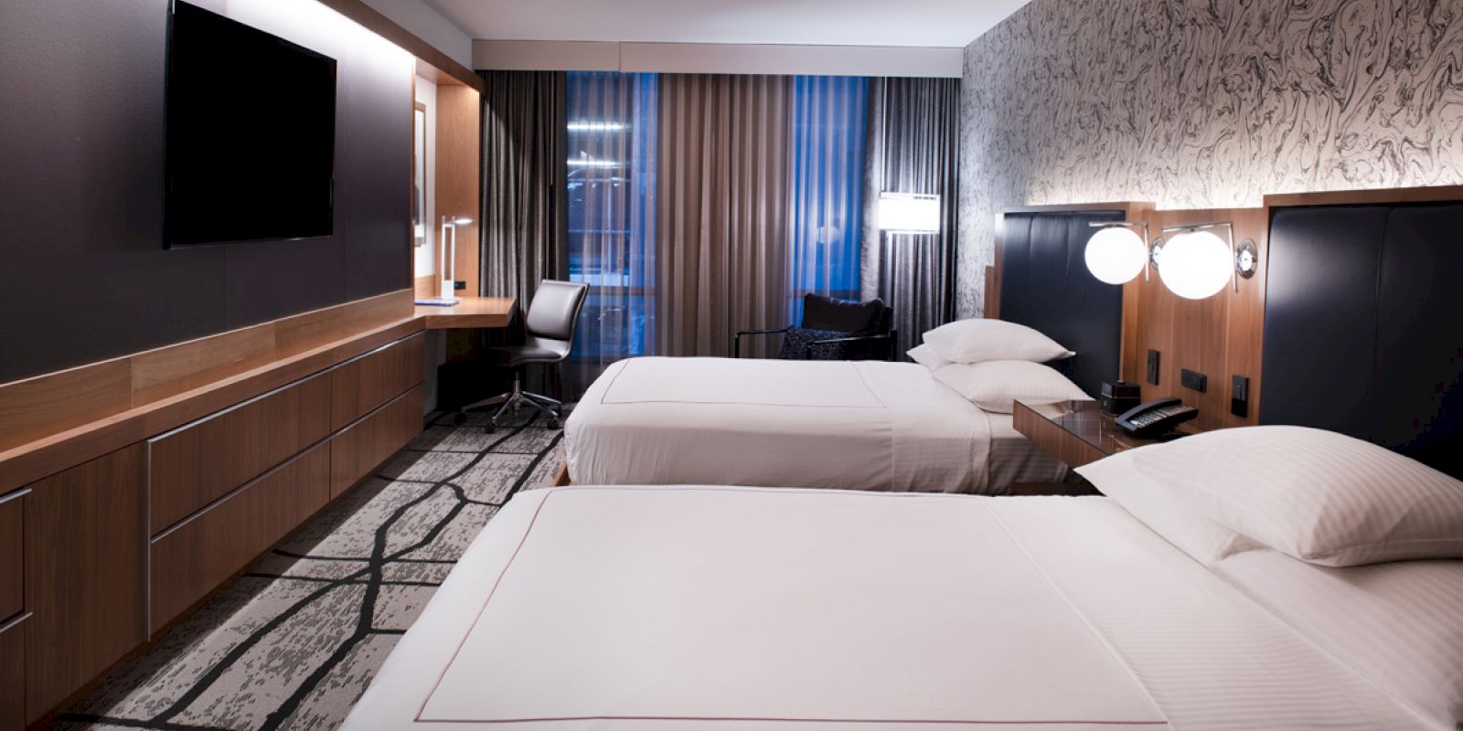 The image shows a modern hotel room with two double beds, a desk, a chair, a TV, and a window with curtains.