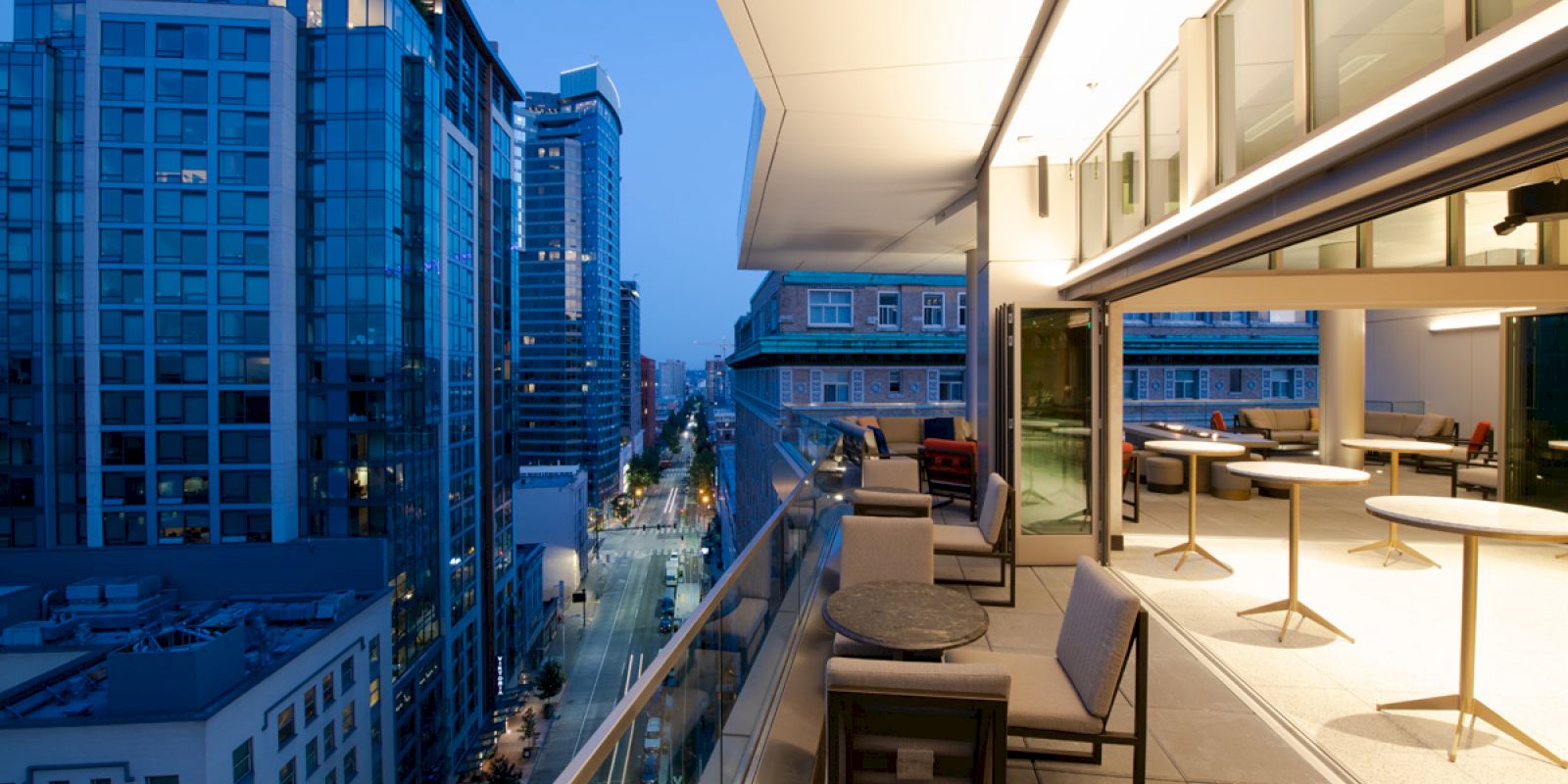 A city view at dusk from a modern rooftop terrace with outdoor seating and tables, overlooking street and tall buildings.