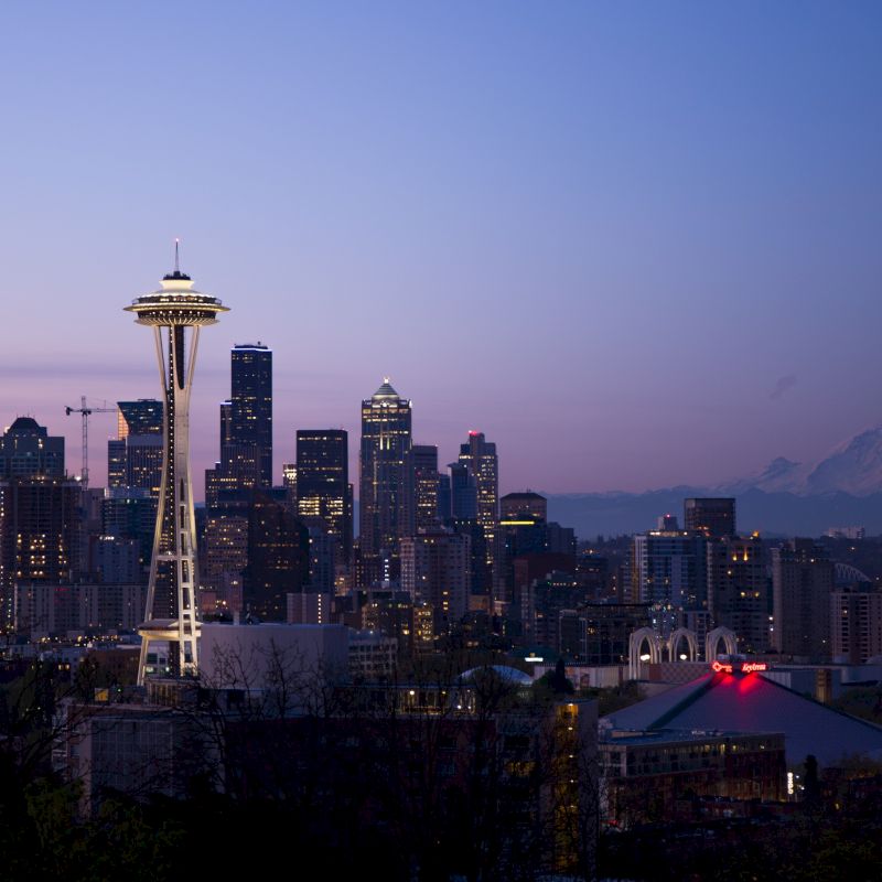 A twilight view of a cityscape featuring the Space Needle in the foreground, with skyscrapers, and a mountain in the background.