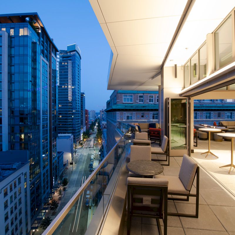 The image shows a modern, high-rise balcony overlooking a city street with tall buildings, featuring outdoor seating and tables under soft evening light.