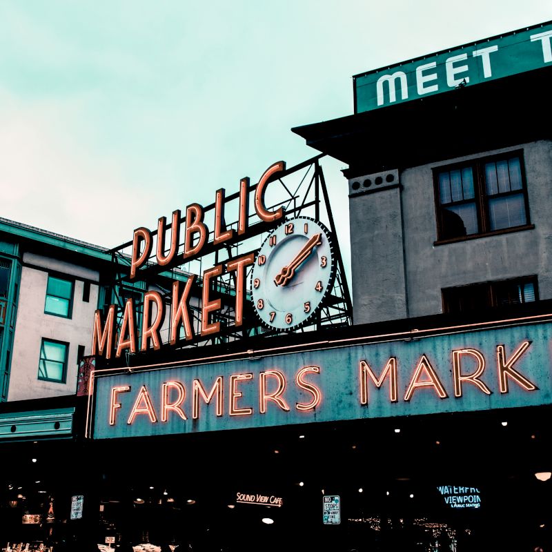 The image features a neon-lit sign for a Public Market and Farmers Market, with an iconic clock, against the backdrop of city buildings.