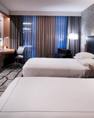 A modern hotel room with two beds, a wall-mounted TV, a work desk by the window, and stylish decor, featuring a patterned wall and contemporary lighting fixtures.