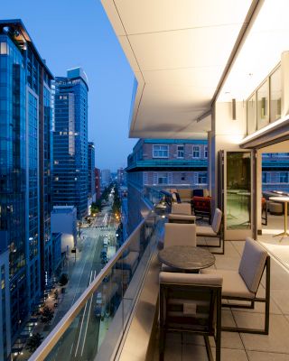 An urban balcony with seating overlooks a city street surrounded by high-rise buildings, lit up as evening begins to set in.