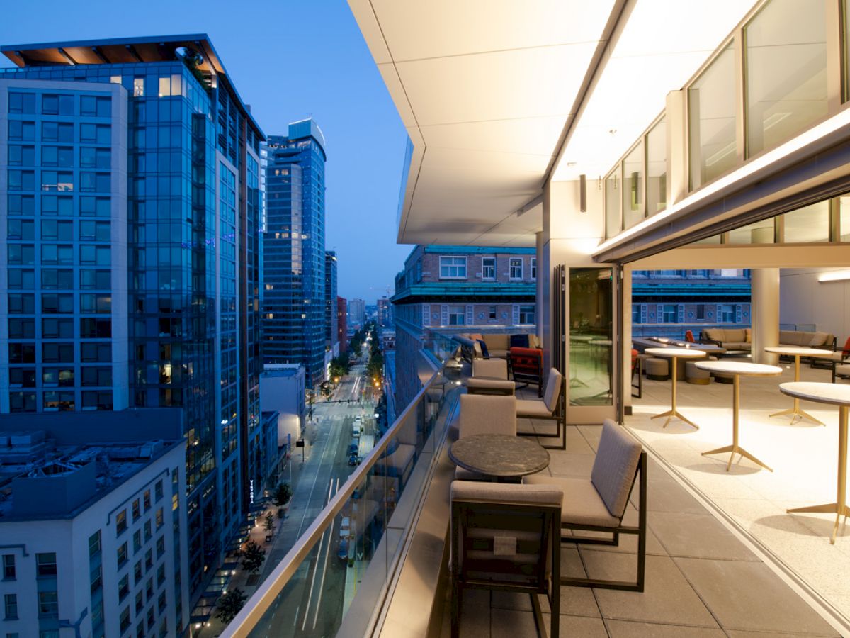 The image shows a modern balcony with seating and tables, overlooking a cityscape with tall buildings and a street lit up in the evening.