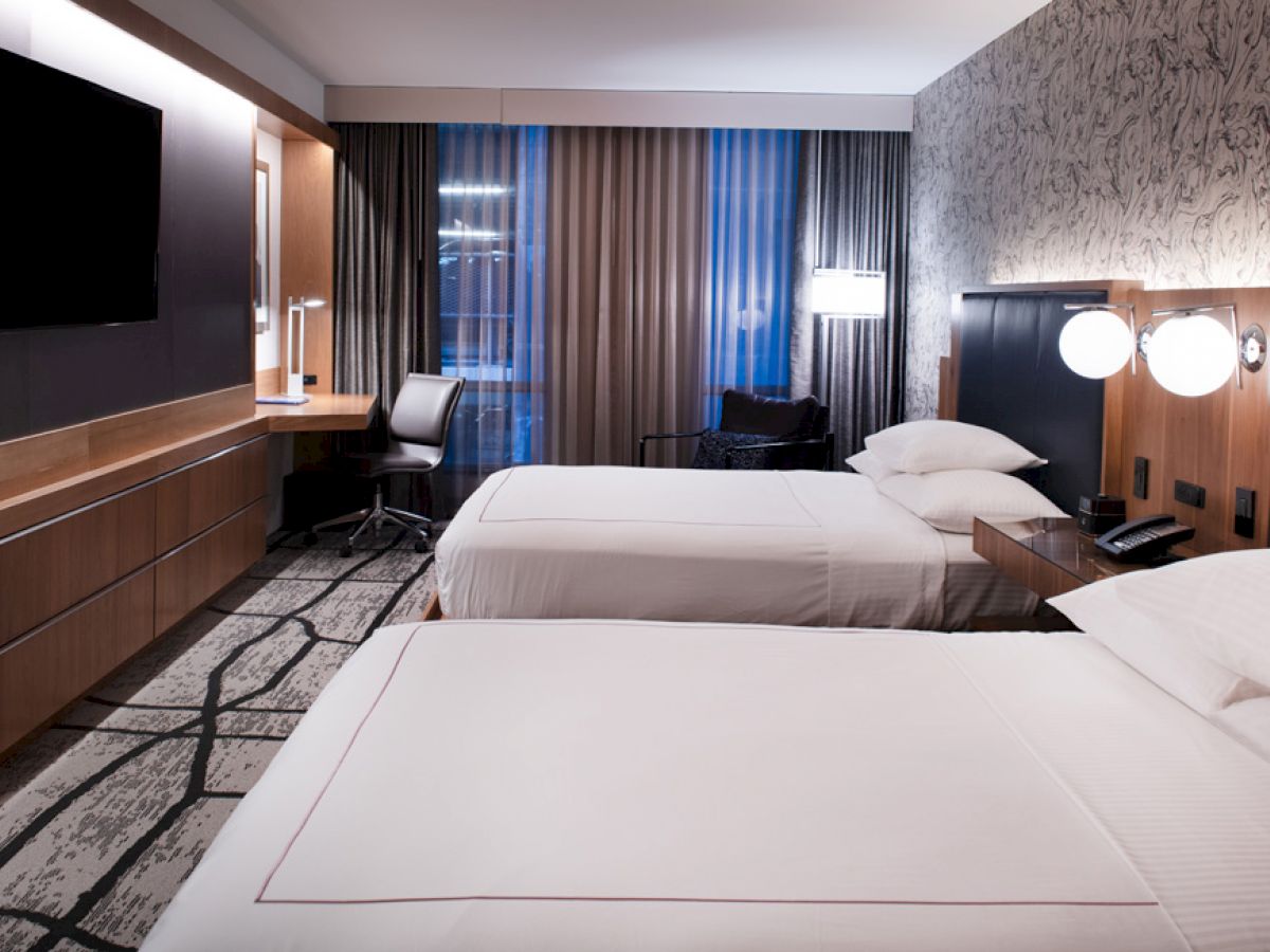A modern hotel room with two beds, a flat-screen TV, a desk, chairs, bedside lamps, a patterned wall, and a large window with curtains.