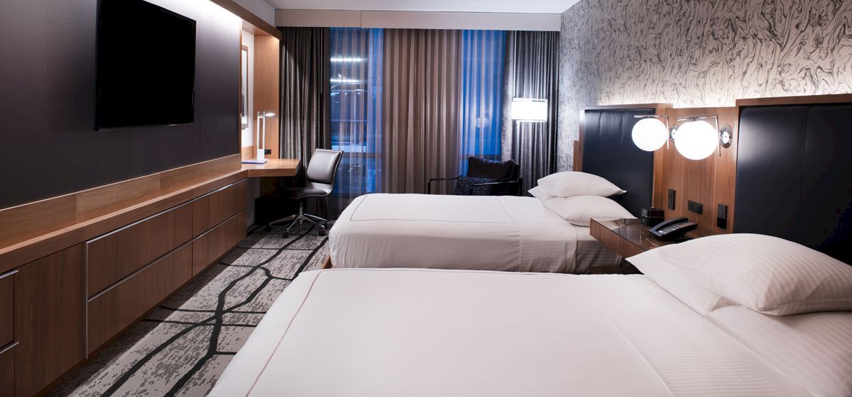 This image shows a modern hotel room with two neatly made beds, a flat-screen TV, a work desk with a chair, and a well-lit, stylish ambiance.