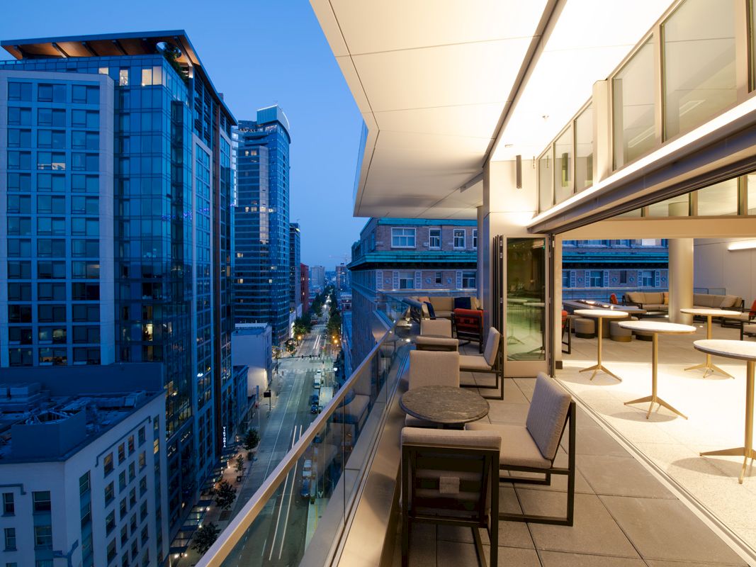 The image shows a modern outdoor terrace overlooking a cityscape at dusk, with buildings lining a street and tables and chairs on the terrace.