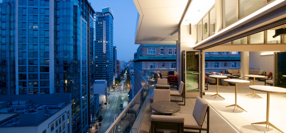 The image shows a modern outdoor terrace overlooking a cityscape at dusk, with buildings lining a street and tables and chairs on the terrace.