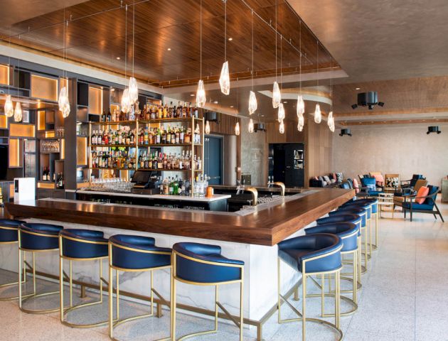 The image shows a modern bar with high shelves of liquor, bar stools, hanging lights, and a lounge area in the background with seating.