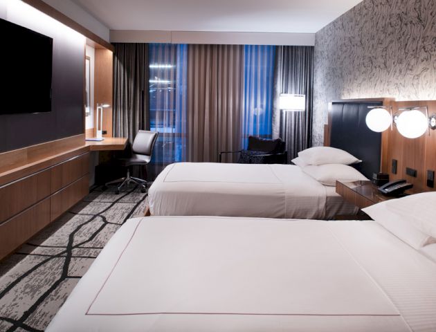The image shows a modern hotel room with two neatly made beds, a flat-screen TV, a desk with a chair, and decorative lighting.