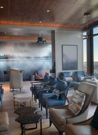 A modern lounge with multiple chairs, tables, large windows offering a scenic view of water and a Ferris wheel, and a mural of a city skyline on the wall.