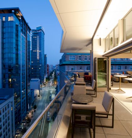 A modern rooftop terrace overlooking a city street, with seating areas and tables, surrounded by tall buildings and city lights at dusk.