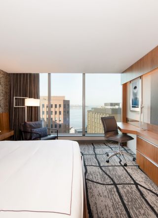 A modern hotel room with a king-sized bed, a wall-mounted TV, a desk, a chair, and a large window showcasing a cityscape view.