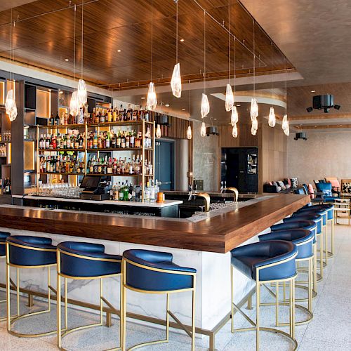 A modern bar with blue high stools, wooden counter, numerous hanging lights, and a well-stocked shelf of bottles; stylish seating area in the background.