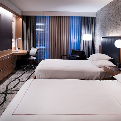 The image shows a modern hotel room with two neatly made beds, a flat-screen TV, a desk, and a chair near large windows with curtains.