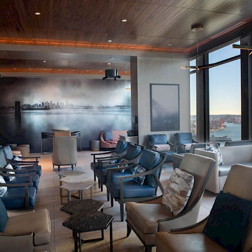 A modern lounge area with numerous chairs, tables, a cityscape mural, and large windows providing a scenic view of a waterfront and Ferris wheel.