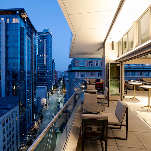 A modern urban balcony with seating and tables, overlooking a city street lined with tall buildings, during the early evening hours.