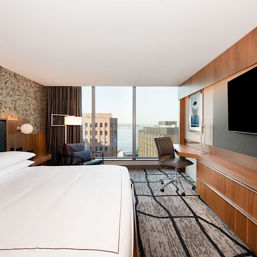 A modern hotel room with a large bed, desk, chair, big-screen TV, and a window offering a city view.