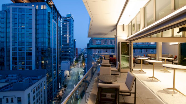 A modern cityscape at twilight featuring tall buildings and a sleek balcony with tables and chairs, providing a scenic view of the urban street below.