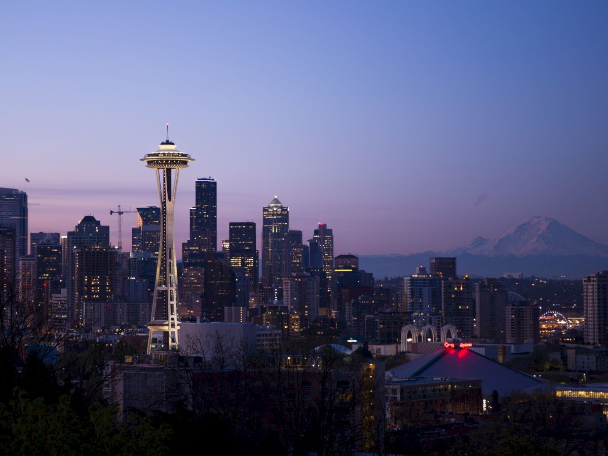 A city skyline at dusk featuring the Space Needle in the foreground and a mountain in the background, under a clear sky.